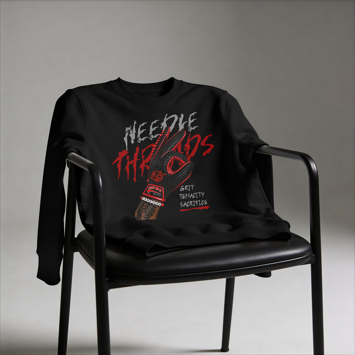 A black crewneck sweatshirt with a bold red, white, and gray graphic print on the front, draped over a black metal chair against a neutral gray studio background.