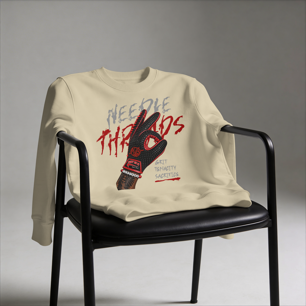 A cream-colored crewneck sweatshirt with a graphic print of a black and red hockey glove and the words 'Needle Threads' and 'Best Trinity Sacrifice', draped over a black chair against a gray studio background.