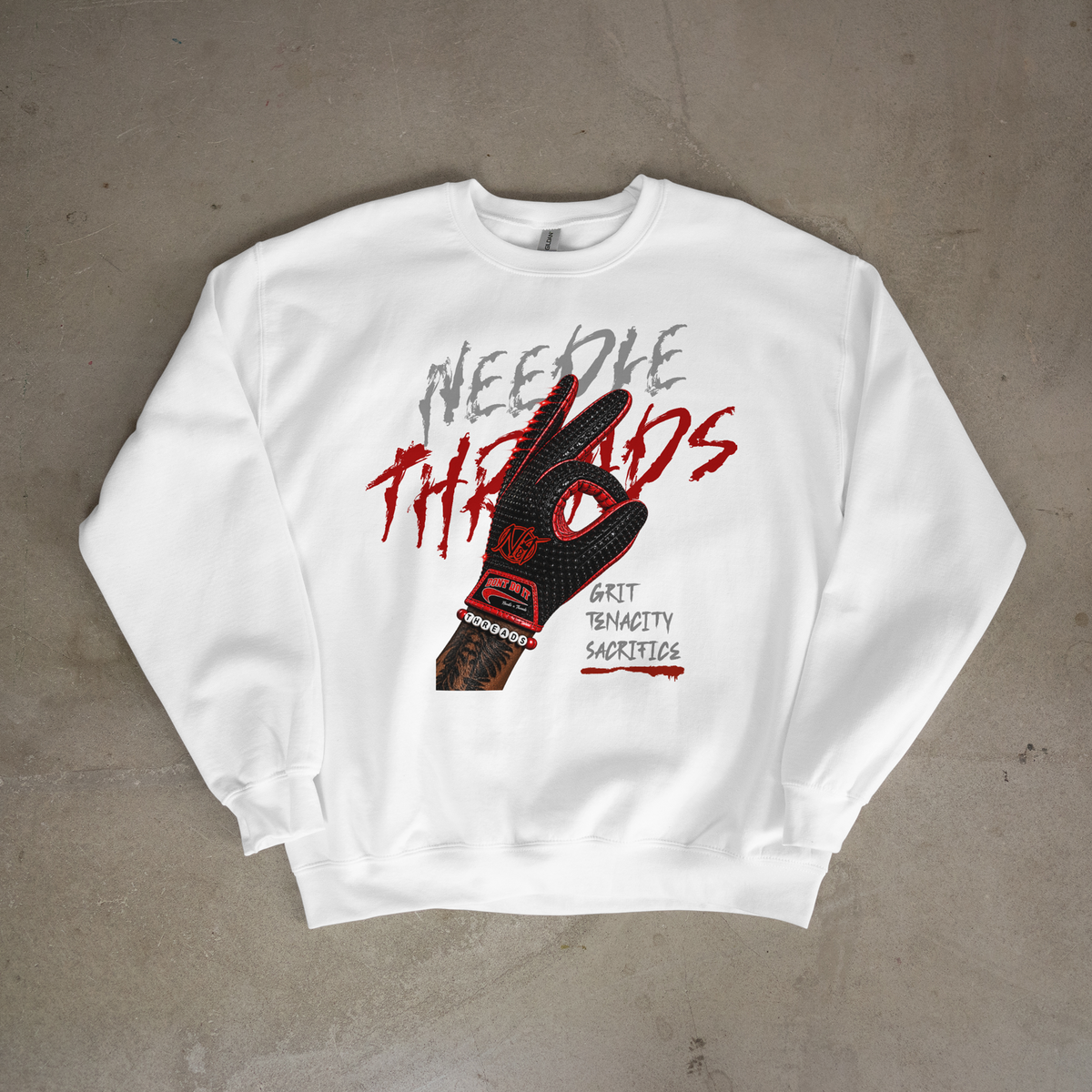 A white crewneck sweatshirt laid flat on a textured gray concrete surface, featuring a graphic print of a black and red baseball glove with the words 'Needle Threads' and 'Grit Tenacity Sacrifice'.