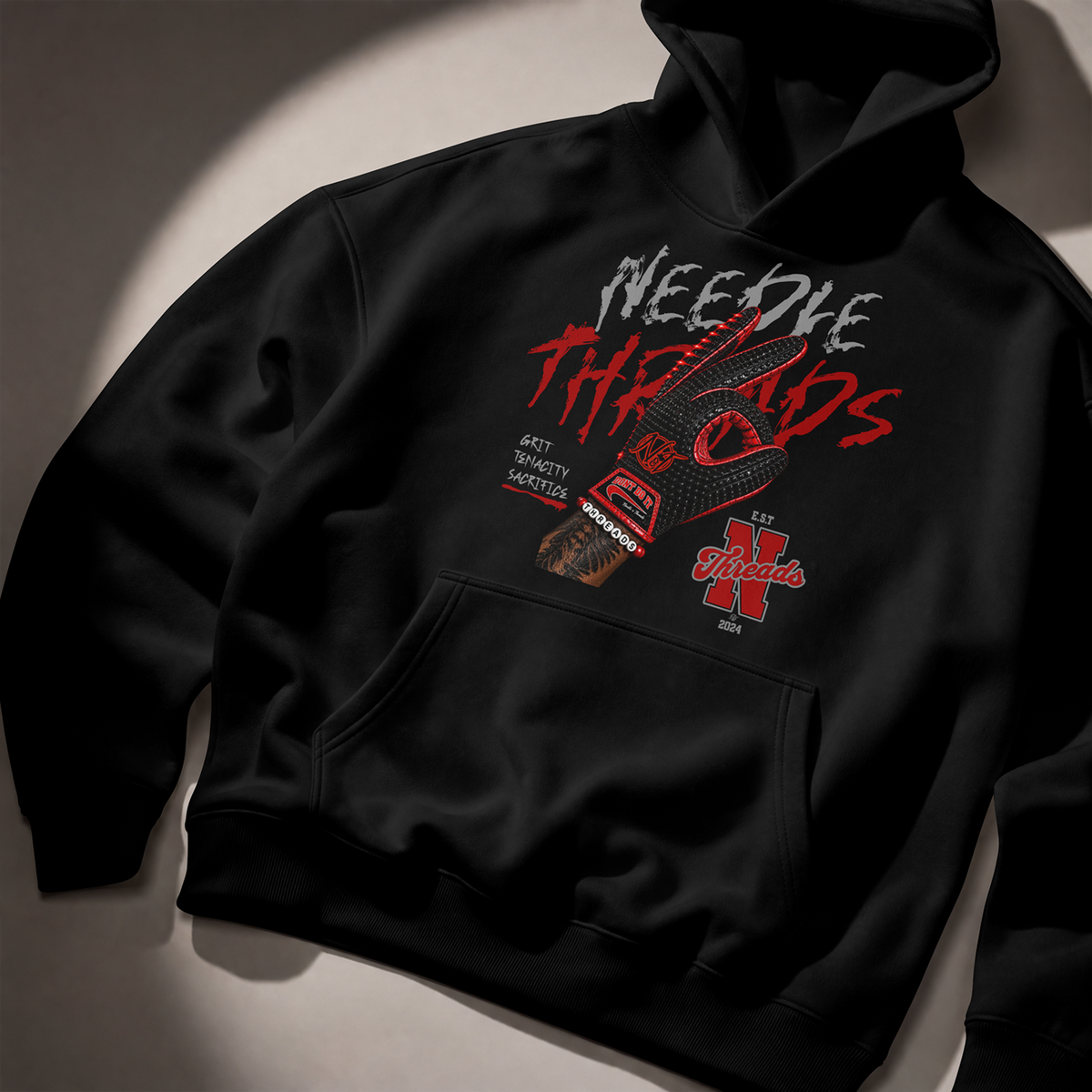 A black pullover hoodie laid flat on a light gray surface with dramatic diagonal lighting. The front has a large distressed graphic reading 'NEEDLE THREADS' in white and red, with a guitar illustration, a baseball bat, and a red stylized 'N' logo.