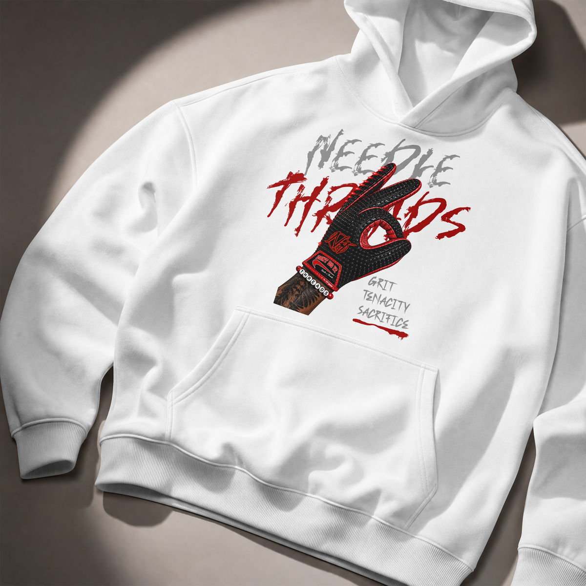 A white pullover hoodie laid flat on a warm beige surface with dramatic diagonal shadows. The front features a large graphic print with the words 'Needle Threads' in gray and red, a black-and-red tattooed hand, and smaller text reading 'Grunt Tenacity Shortage.