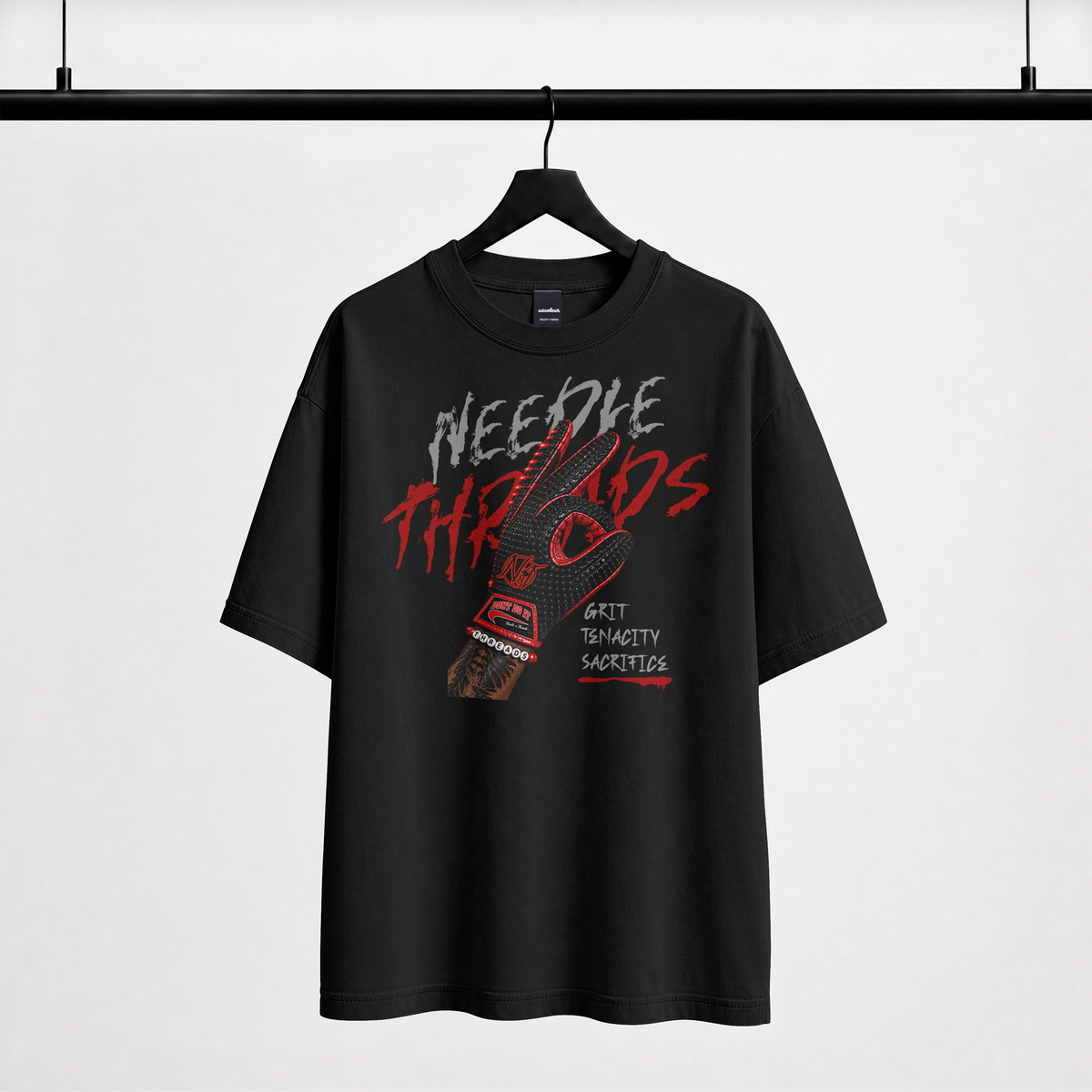 A black short-sleeve graphic T-shirt hanging on a black hanger against a light gray background. The shirt features a large front graphic with the words 'NEEDLE THREADS' in distressed white and red lettering, a red-and-black shoe illustration, and smaller text reading 'GRIT TENACITY SACREDNESS'.