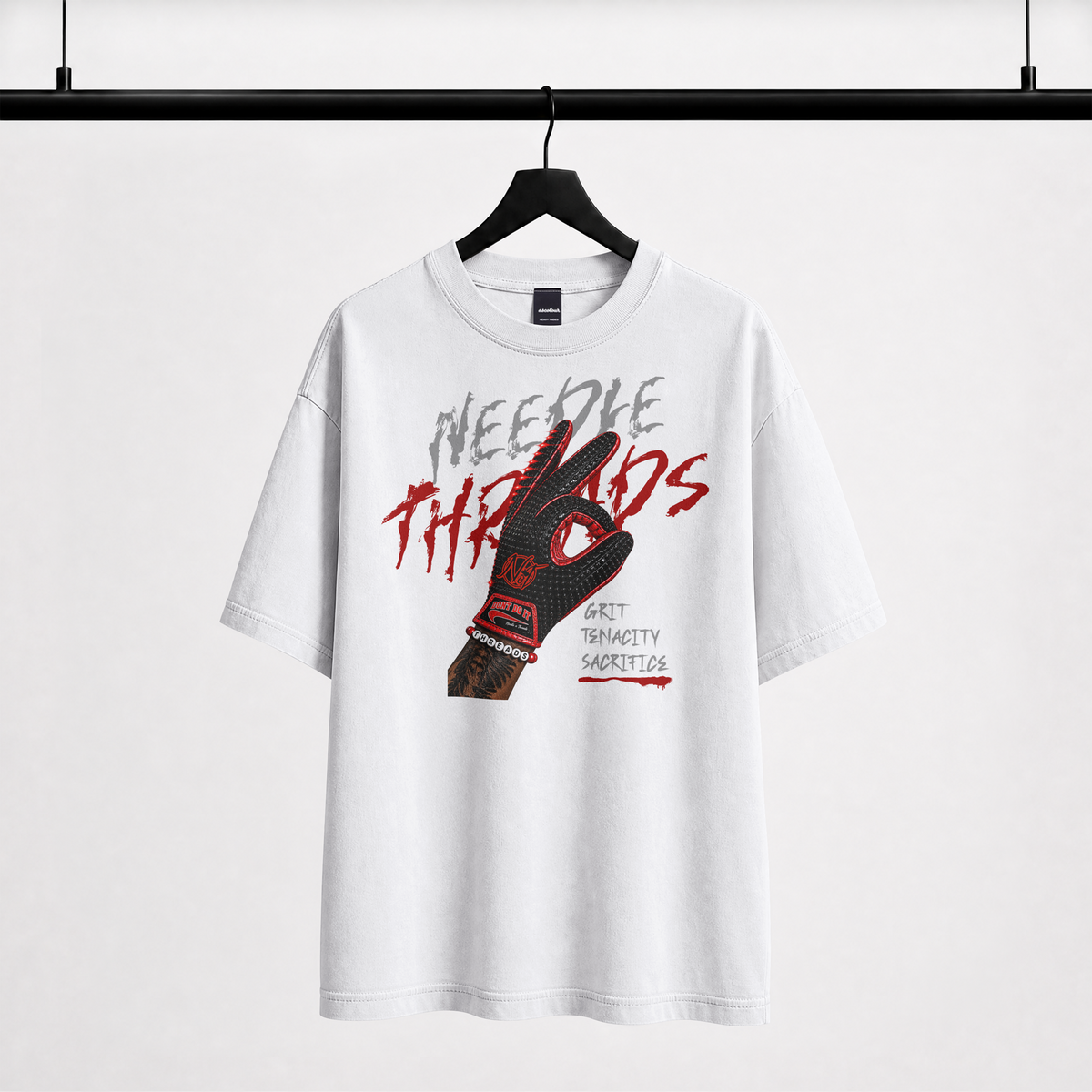 A white short-sleeve graphic T-shirt hanging on a black hanger against a light gray background. The shirt features a large front graphic with the words 'Needle Threads' in gray and red, overlaid on a black-and-red glove illustration, with smaller text reading 'Soft Tragedy Virtuous' and 'Terrorism' on the glove.